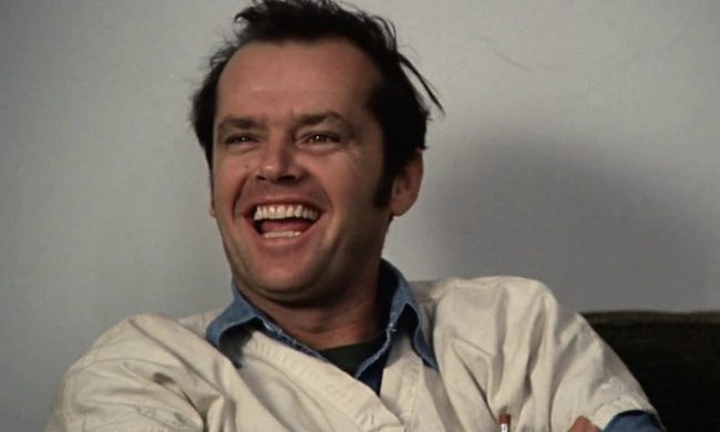 Jack Nicholson sits against a wall with his hands crossed and smiles.